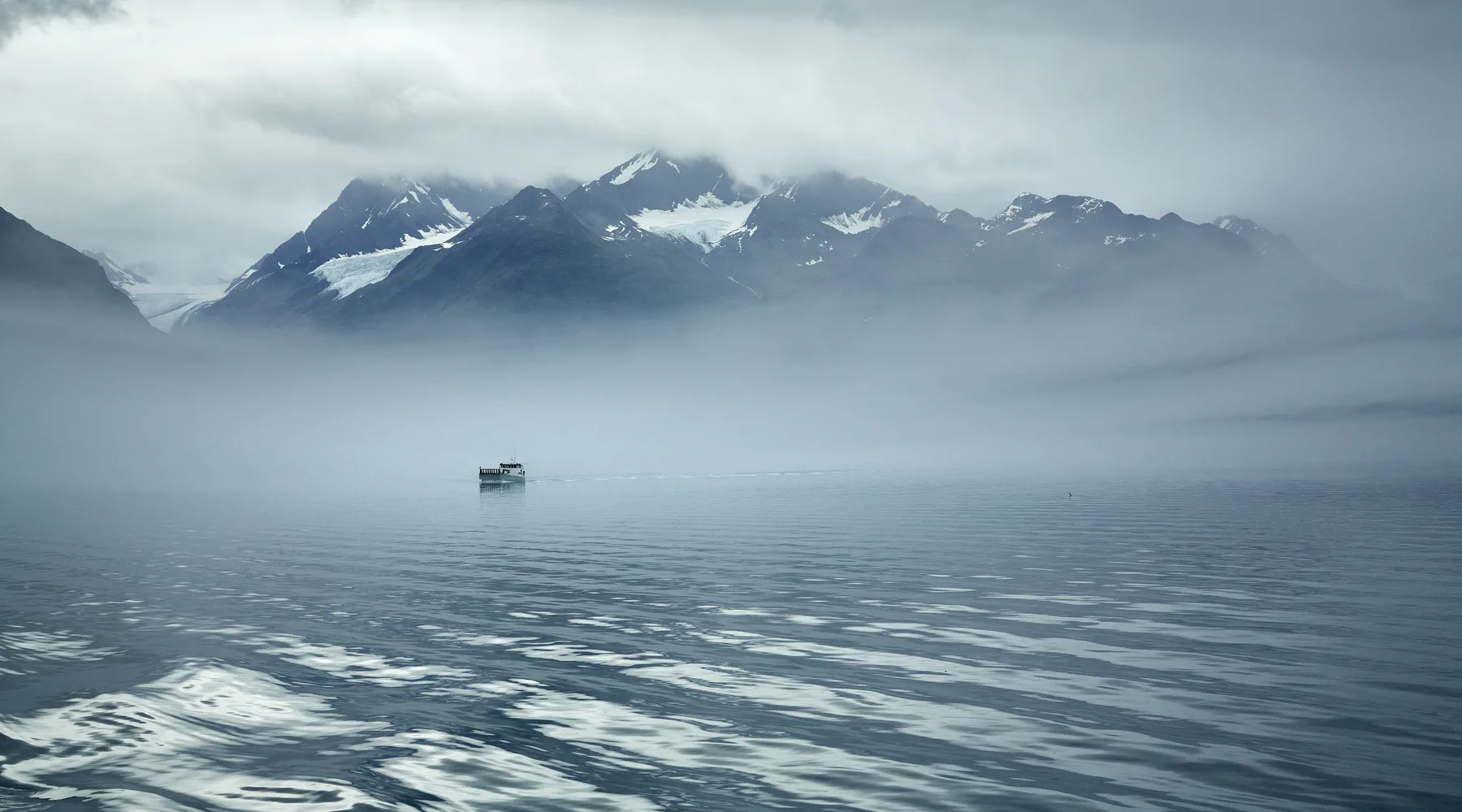 Small boat crossing calm water beneath misty mountains in Valdez, Alaska